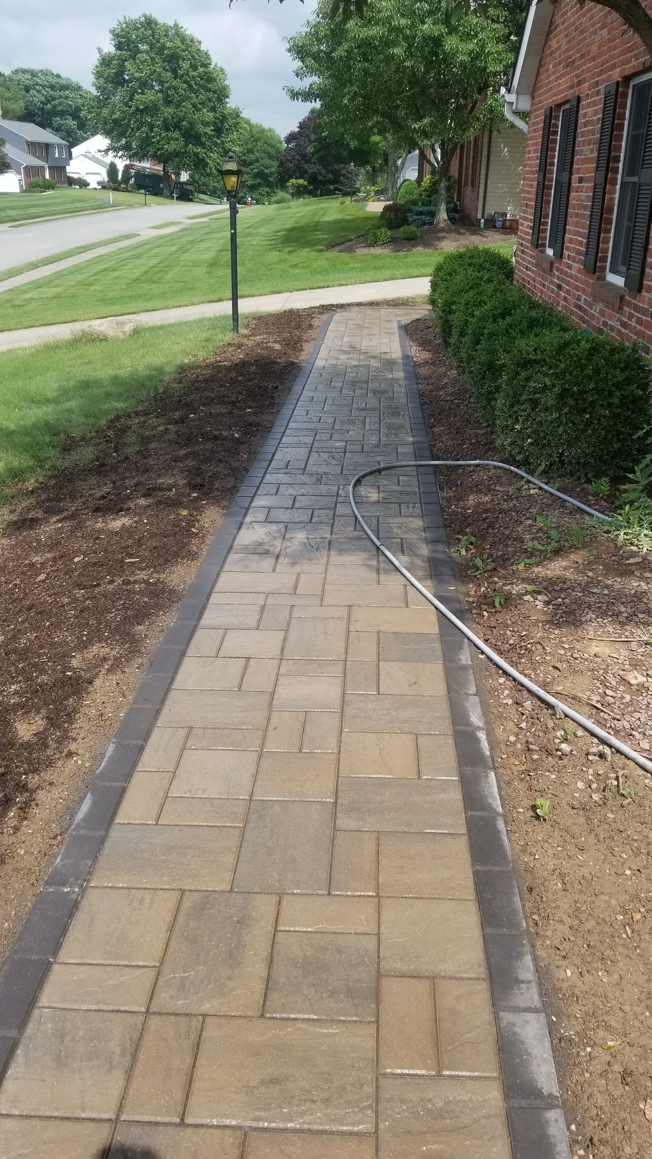 Brick walkway leading to a house, with mulch beds on each side. A hose lies across the path.