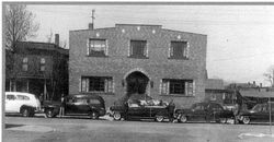 Black-and-white street view of a brick building with parked vintage cars in front.