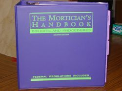 Purple binder titled “The Mortician’s Handbook” on a wooden table.