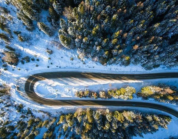 Aerial view of a winding road through a snowy forest with sunlit trees.