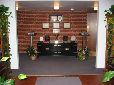 Lobby reception desk against a brick wall, with plants, lamps, and plaques.
