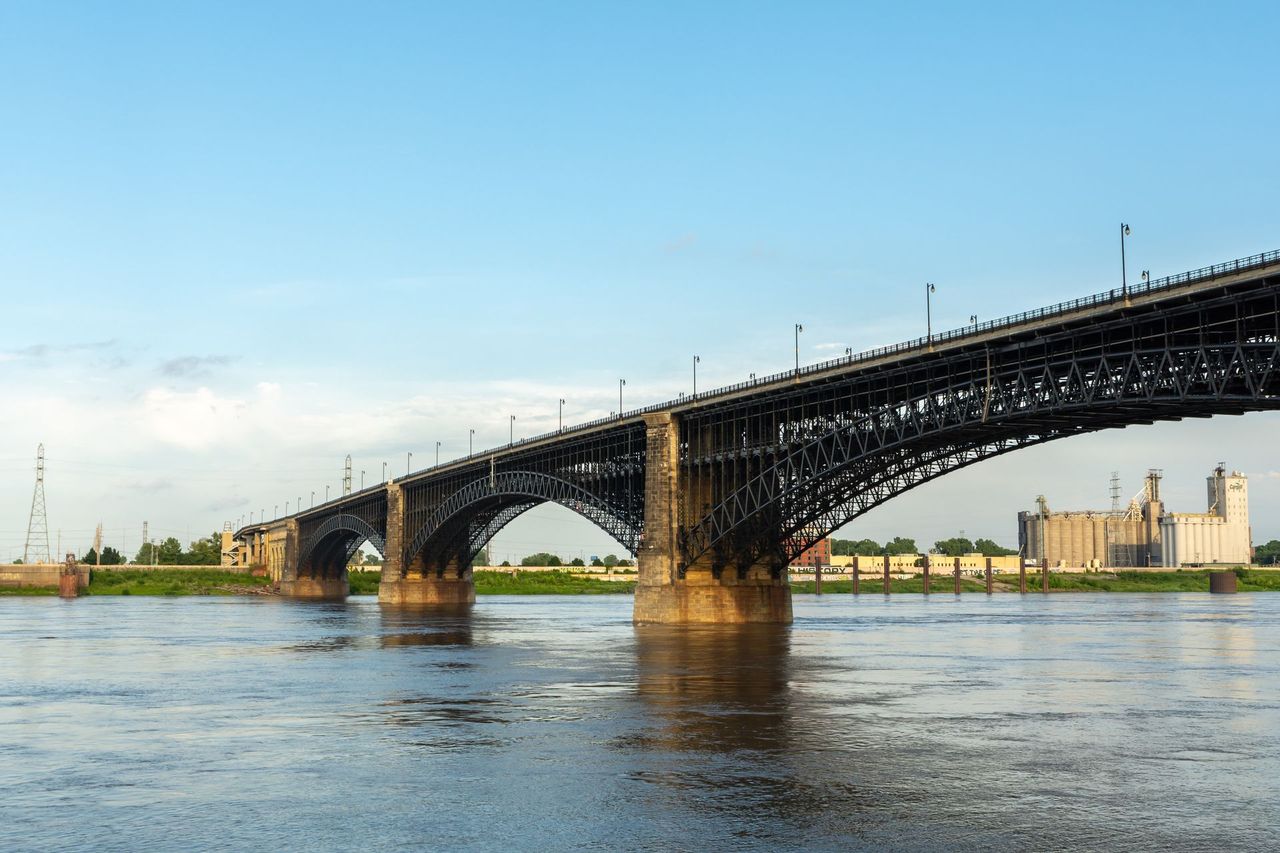 Low bridge over a wide river at sunset, with city buildings in the background