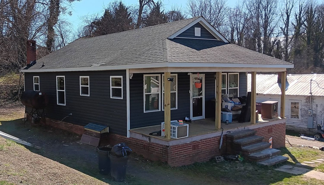 Small dark-gray house with white trim and a brick porch in a wooded yard.