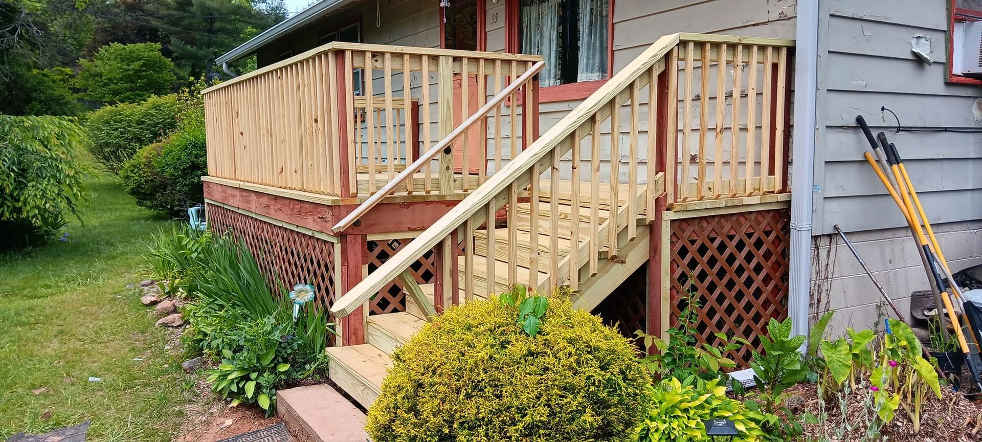 Wooden ramp and porch steps with railing beside a gray house and greenery