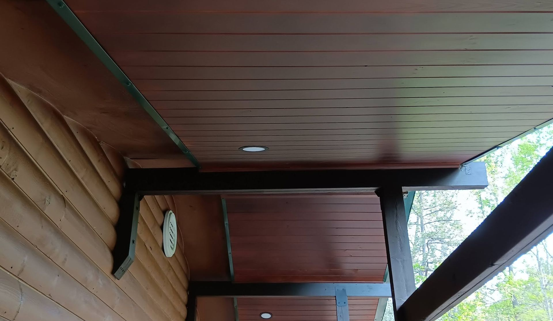 Wooden porch ceiling and wall with beams, viewed from below, with trees visible outside.