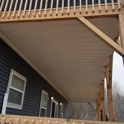 Covered wooden deck under a house roof, with dark siding and railing posts in a wooded area.