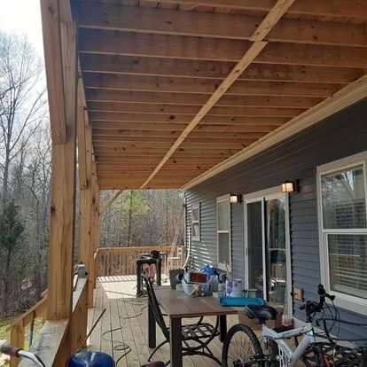 Covered wooden porch with railing, table, bicycle, and sliding doors beside a gray house overlooking trees