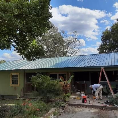 Worker pouring concrete beside a green house with a blue roof and trees overhead