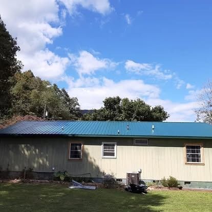 Single-story cream house with a teal roof, set against trees and a blue sky with clouds.