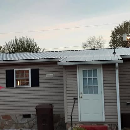 Single-story house exterior with gray siding, white door, black shutters, and a metal roof at dusk