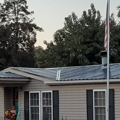 Single-story house with gray siding and a metal roof, trees behind, and an American flag on a pole in front.
