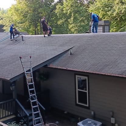 Workers repairing a house roof, with a ladder leaning against the eaves and trees in the background