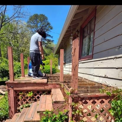 Person standing on a damaged wooden porch beside a house with broken steps and railing.