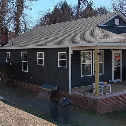 Dark gray house with brick foundation, small porch, and shingled roof on a sunny day