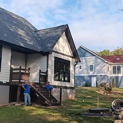 Two workers repairing a house exterior beside stairs, with two neighboring homes in the background.