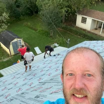 People roofing a house on a steep roof; man taking a selfie in the foreground, backyard and shed below.