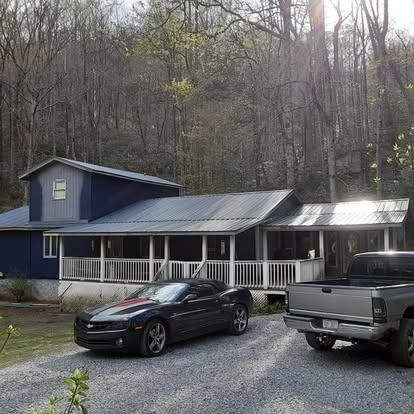 Blue house with a white porch in a wooded area, with a black car and gray pickup in the driveway
