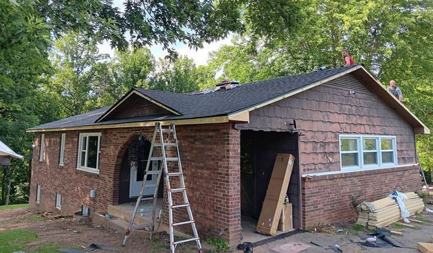 Brick house under repair with ladder, open garage, and exposed roof framing in a wooded yard