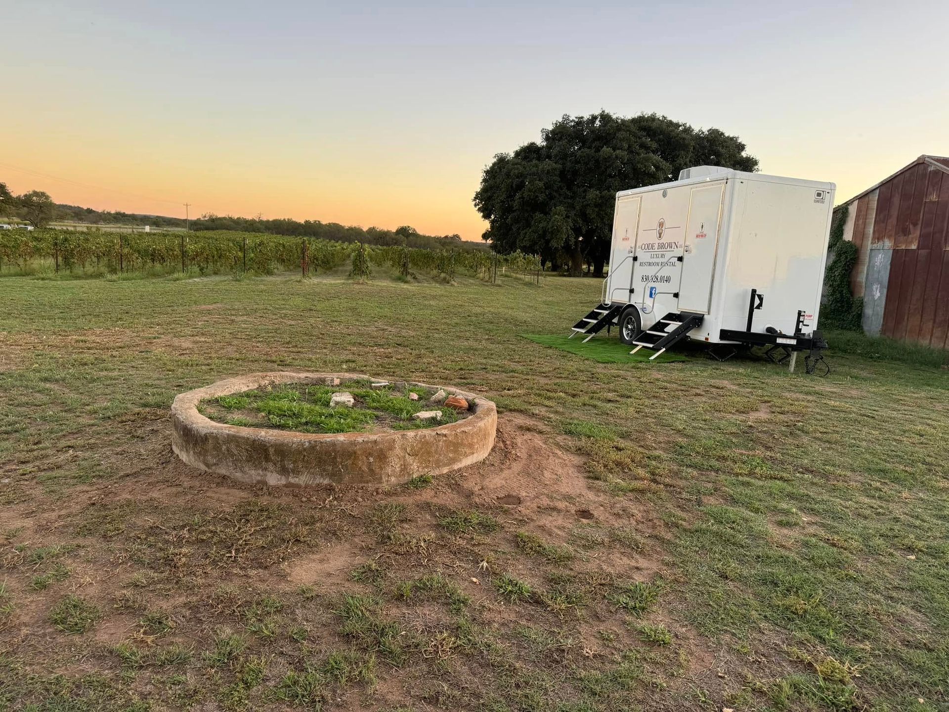 Grassy field at sunset with a small circular fire pit and a white trailer near a brick building.