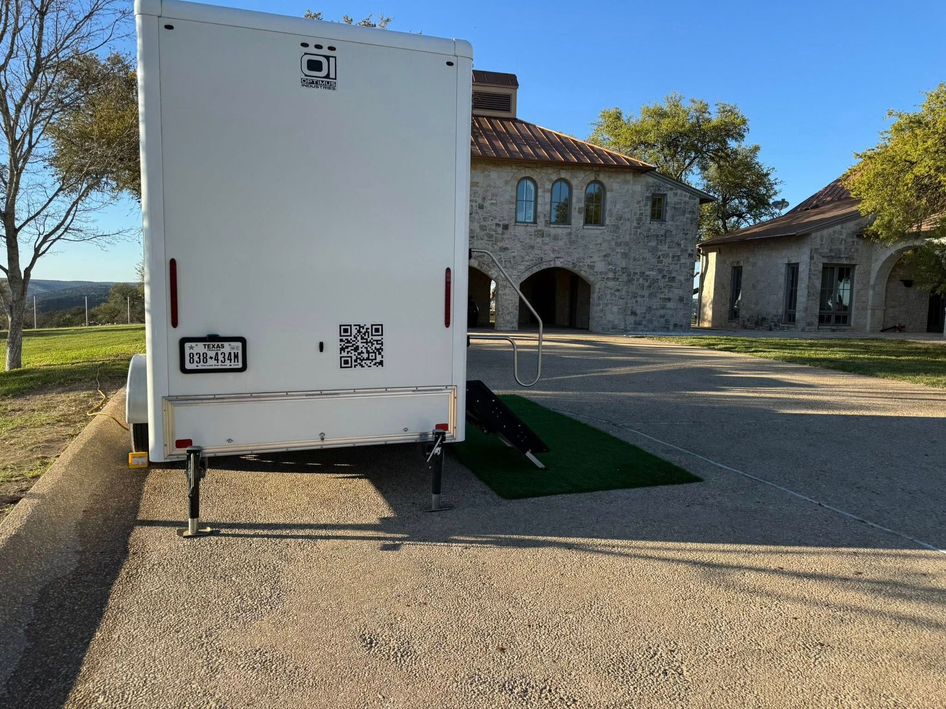 White trailer parked on a gravel lot beside stone buildings, with a small ramp and trees in the background