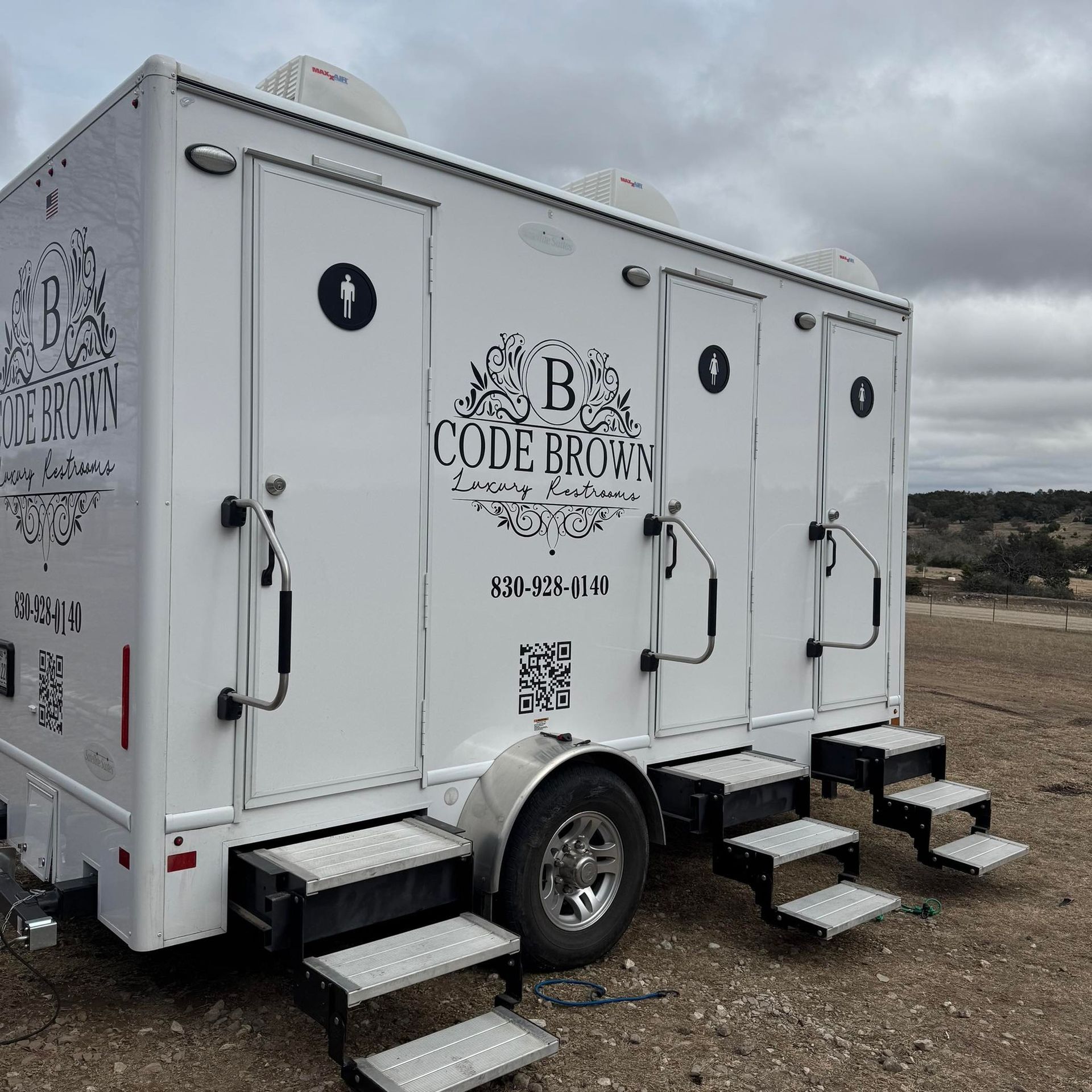White mobile restroom trailer with stairs, parked on gravel under a cloudy sky.