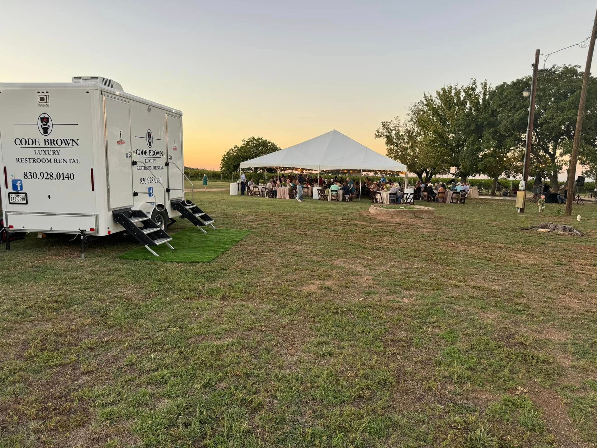 Outdoor event setup on a grassy field with a white tent, trailer, and people gathering at sunset.