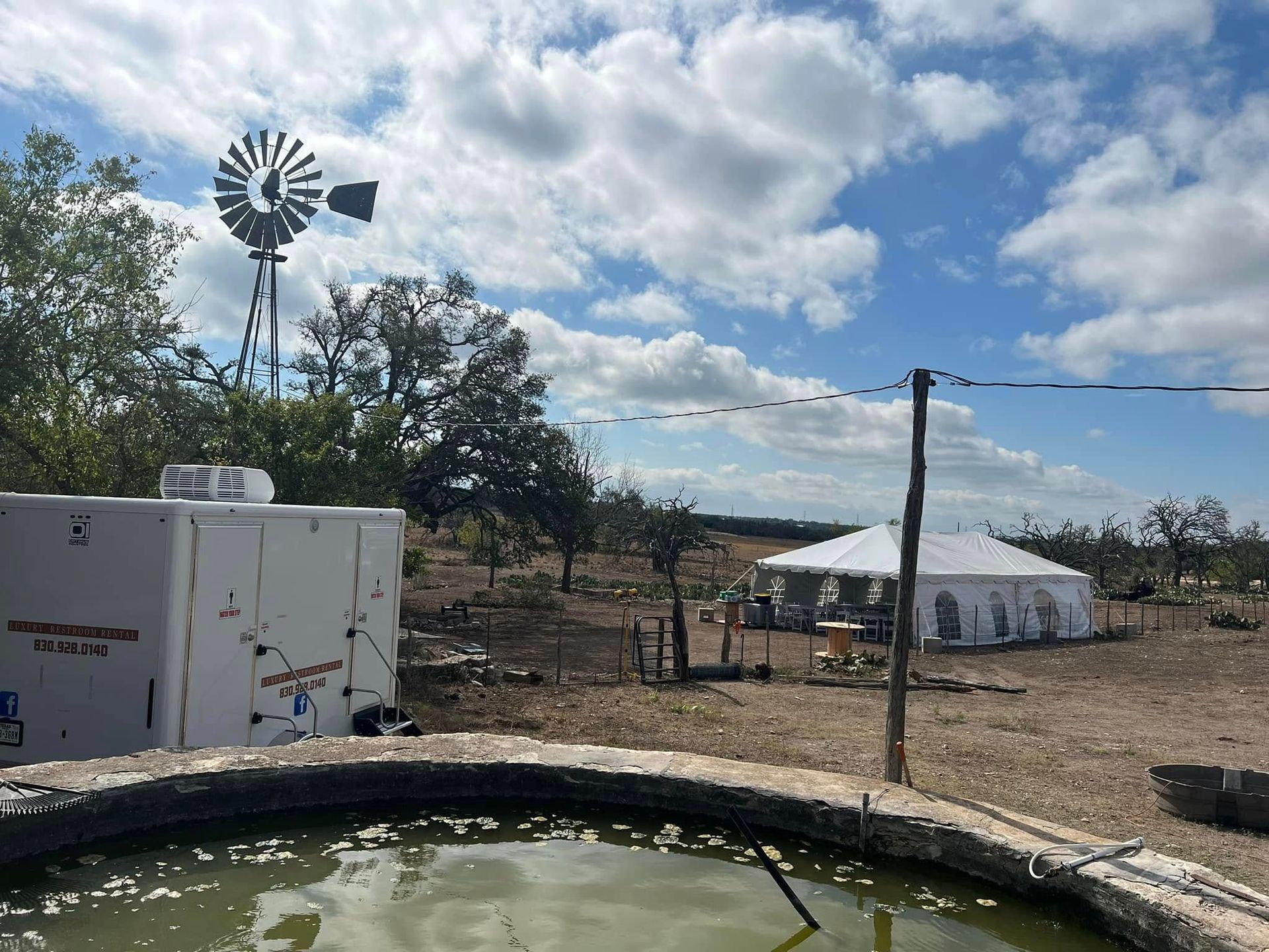 Rural yard with a windmill, white tent, utility box, and a round water tank under a cloudy sky