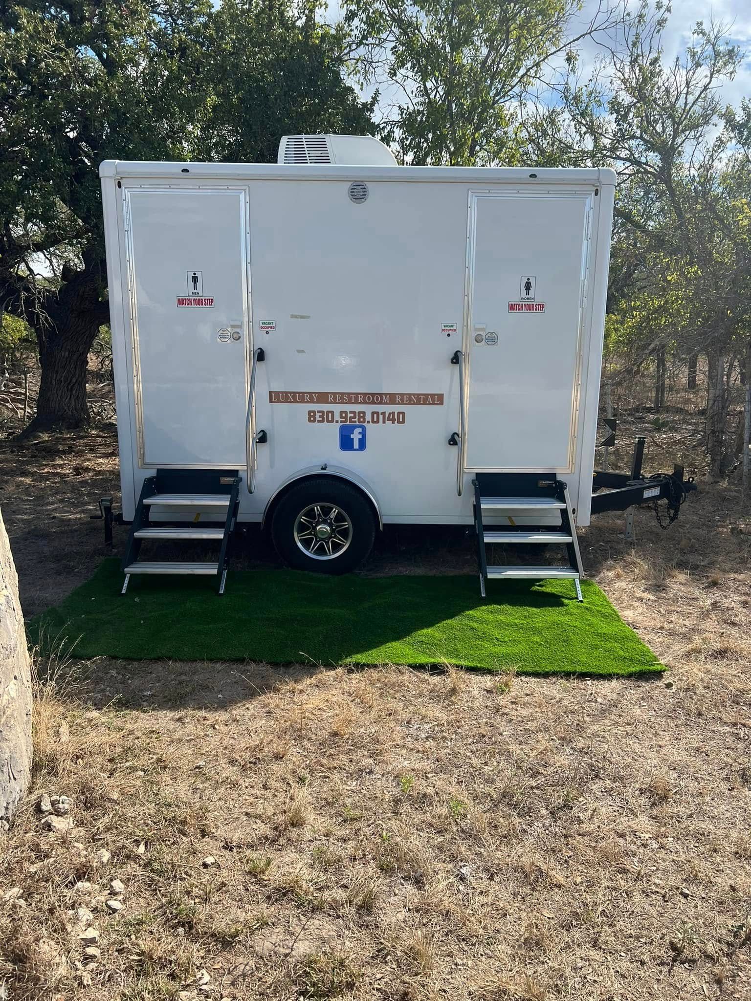 White portable restroom trailer with steps, parked on grass in a wooded outdoor area.