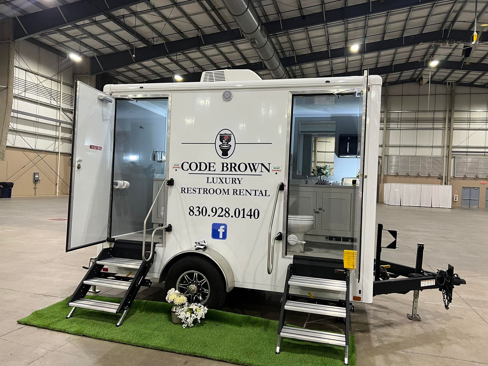 White mobile restroom trailer with flower boxes, stairs, and branding, parked inside a warehouse.
