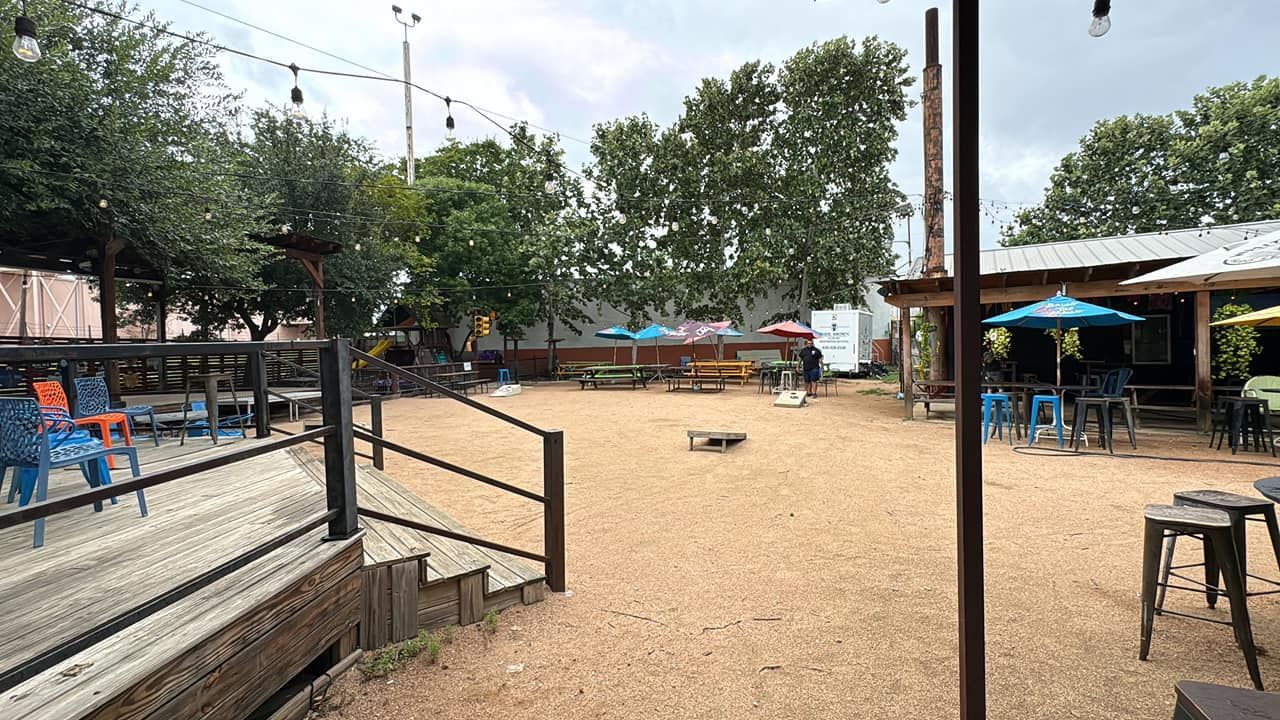 Outdoor gravel courtyard with scattered stools, trees, and small market-style stalls under a cloudy sky