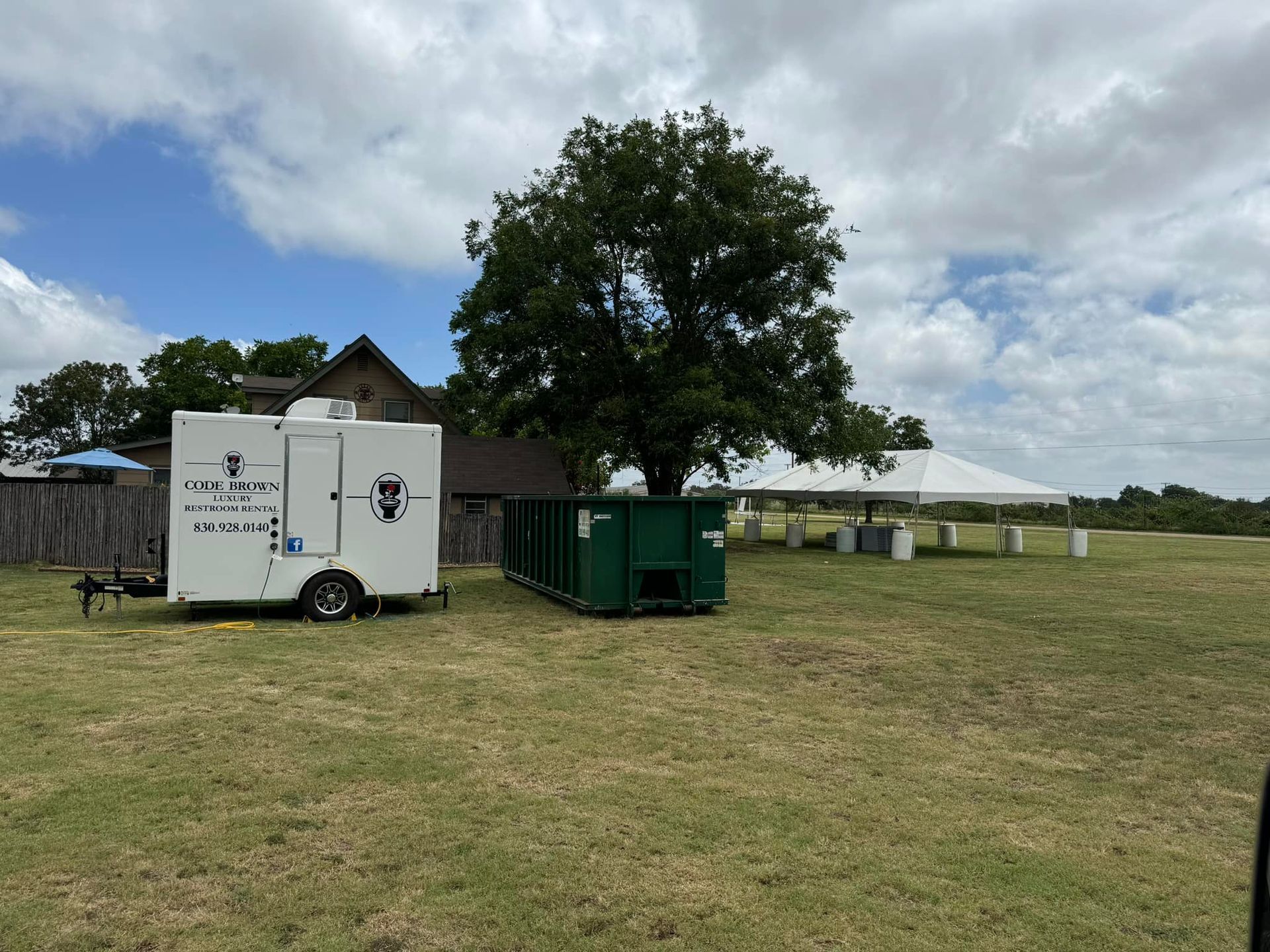 Portable toilets and green dumpsters on a grassy field beside a trailer, under a cloudy sky