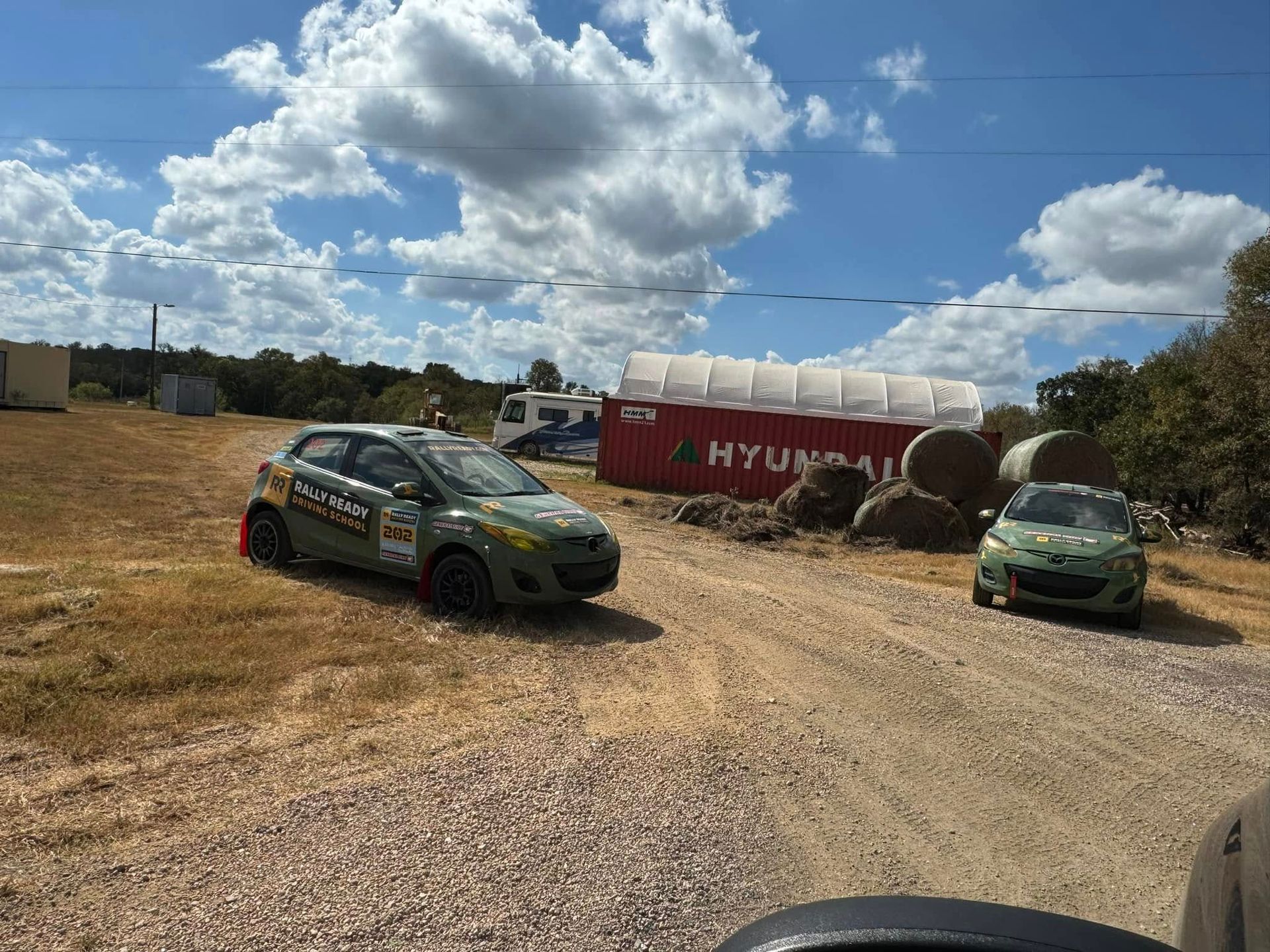 Two rally cars on a dirt track beside a red Hyundai shipping container under a cloudy sky