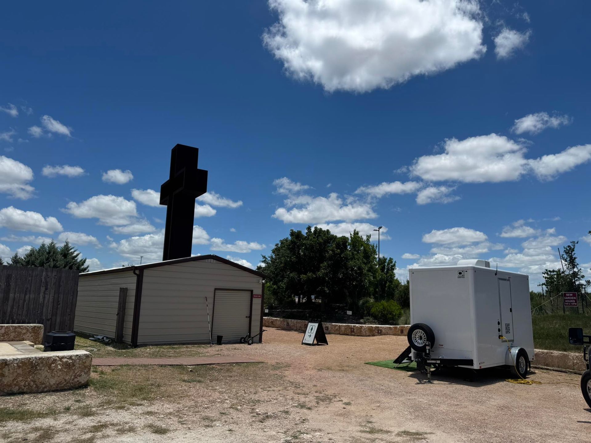 Large black cross beside a beige shed under a bright blue sky with clouds, with two trailers in a dirt lot