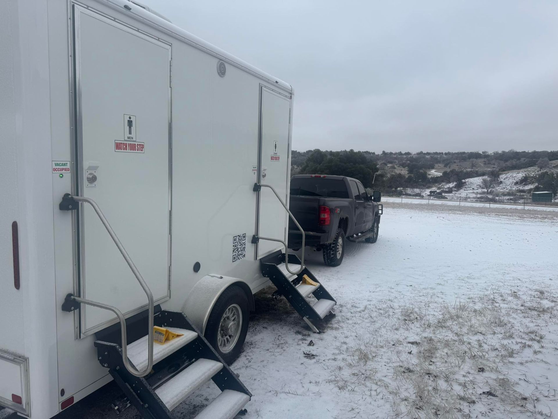 White cargo trailer and black pickup truck parked on snowy ground by a riverbank, with a small step ladder.