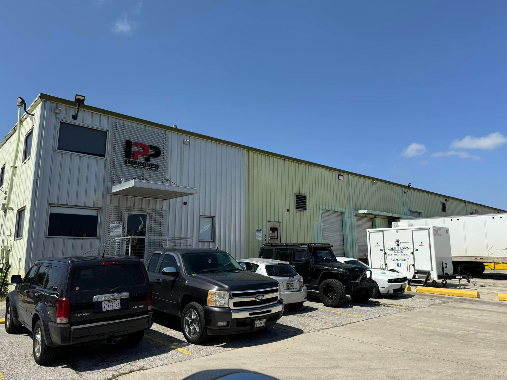 Industrial warehouse with several parked vehicles and a white trailer under a blue sky