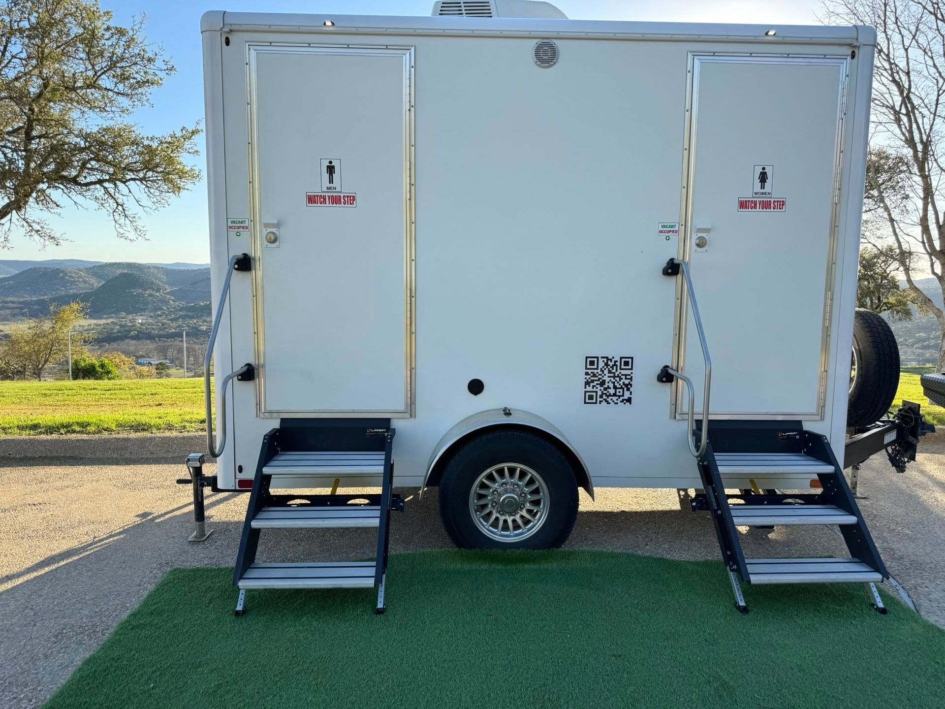 White portable restroom trailer with two entrances and fold-out steps parked outdoors on a roadside.