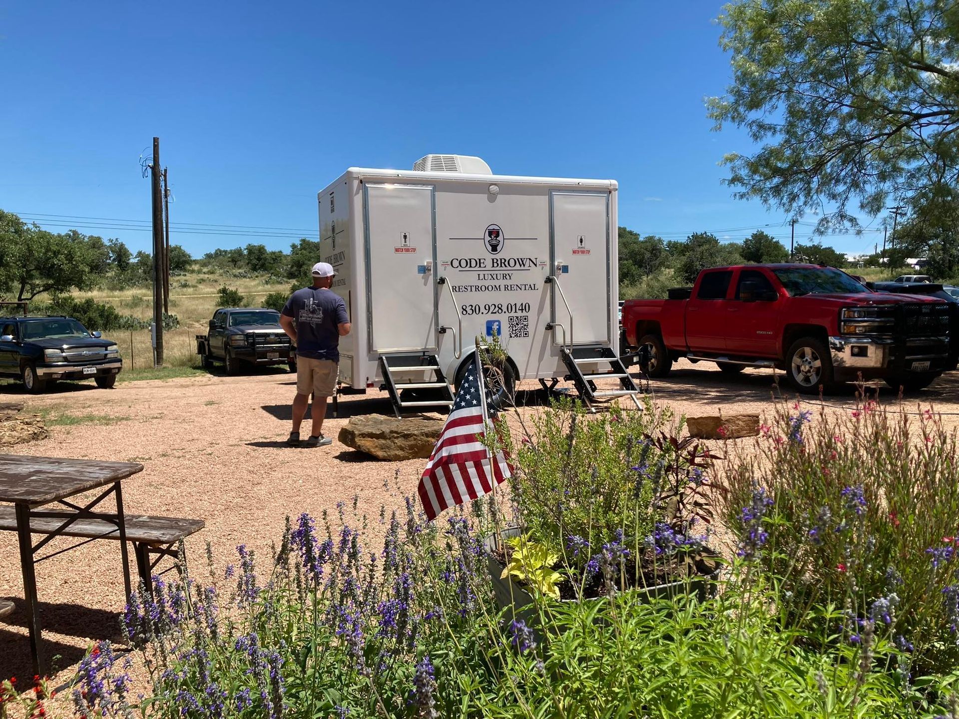 White trailer in a desert campsite with a person standing nearby, red truck parked beside it, and an American flag in front.