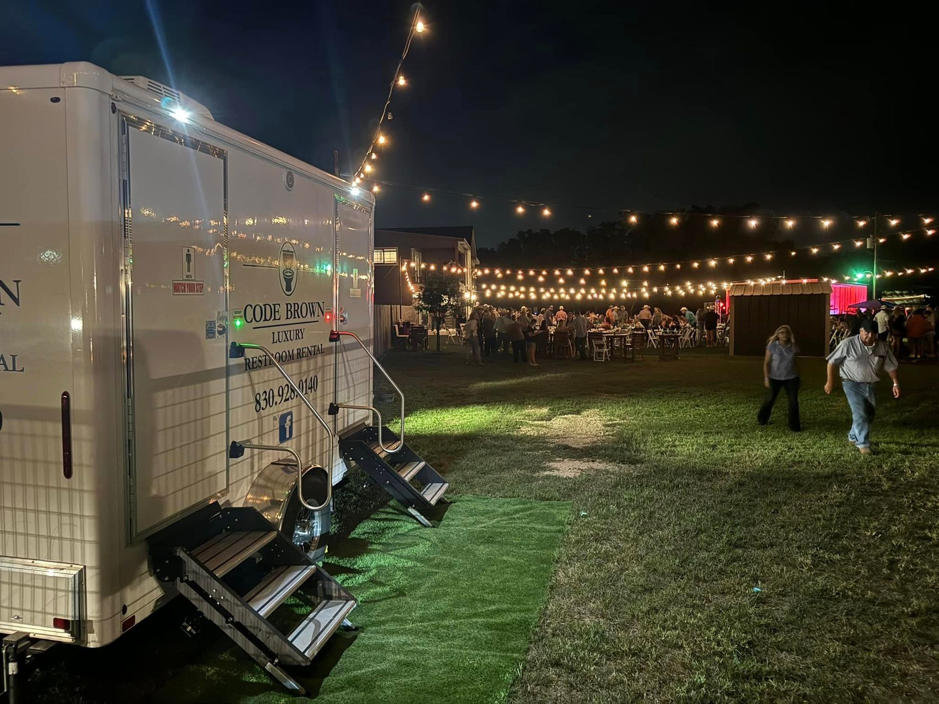 String-lit outdoor event beside a white trailer, with people walking on grass at night.