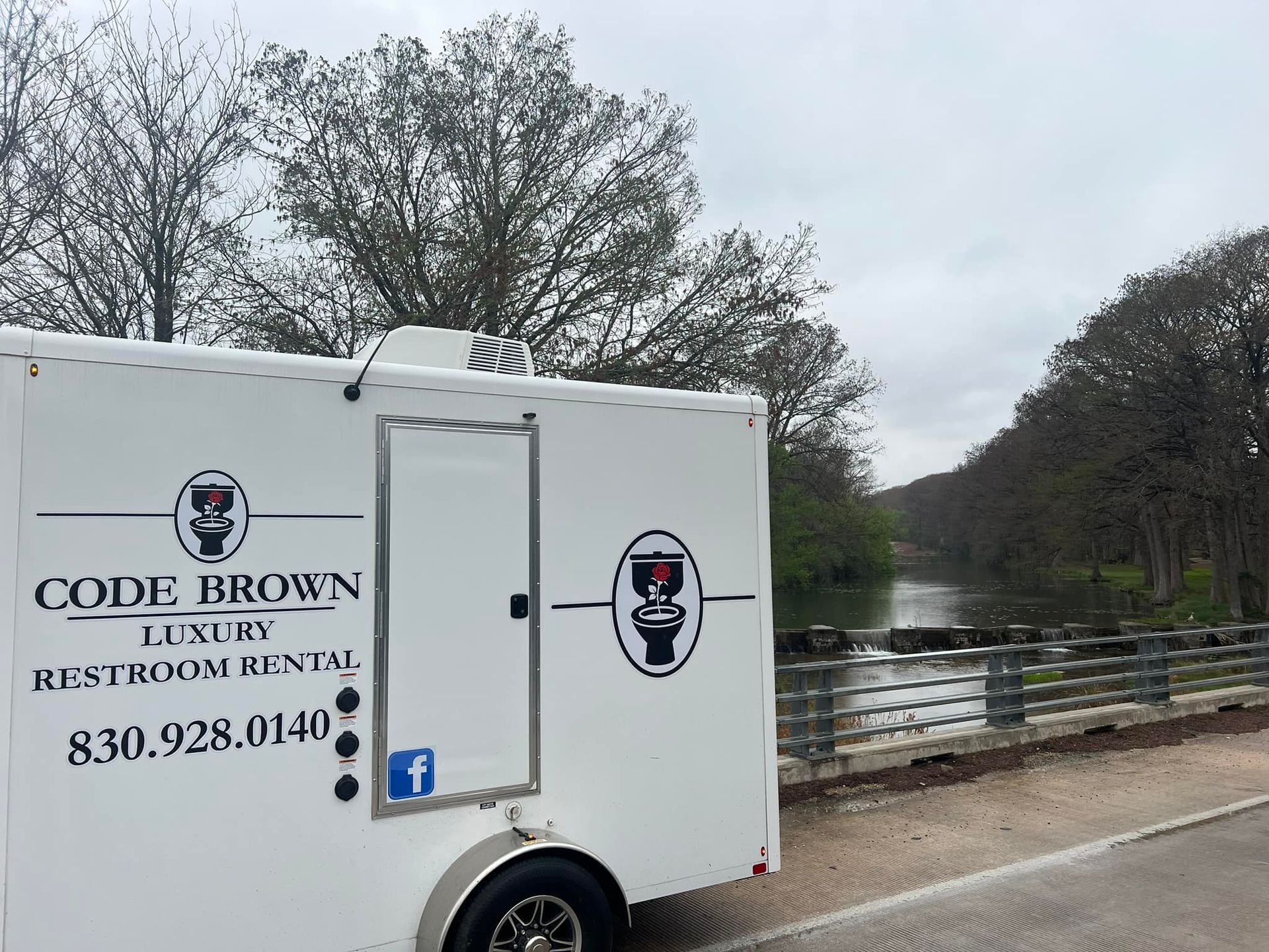 White restroom rental trailer by a riverside bridge, with trees and cloudy sky in the background.