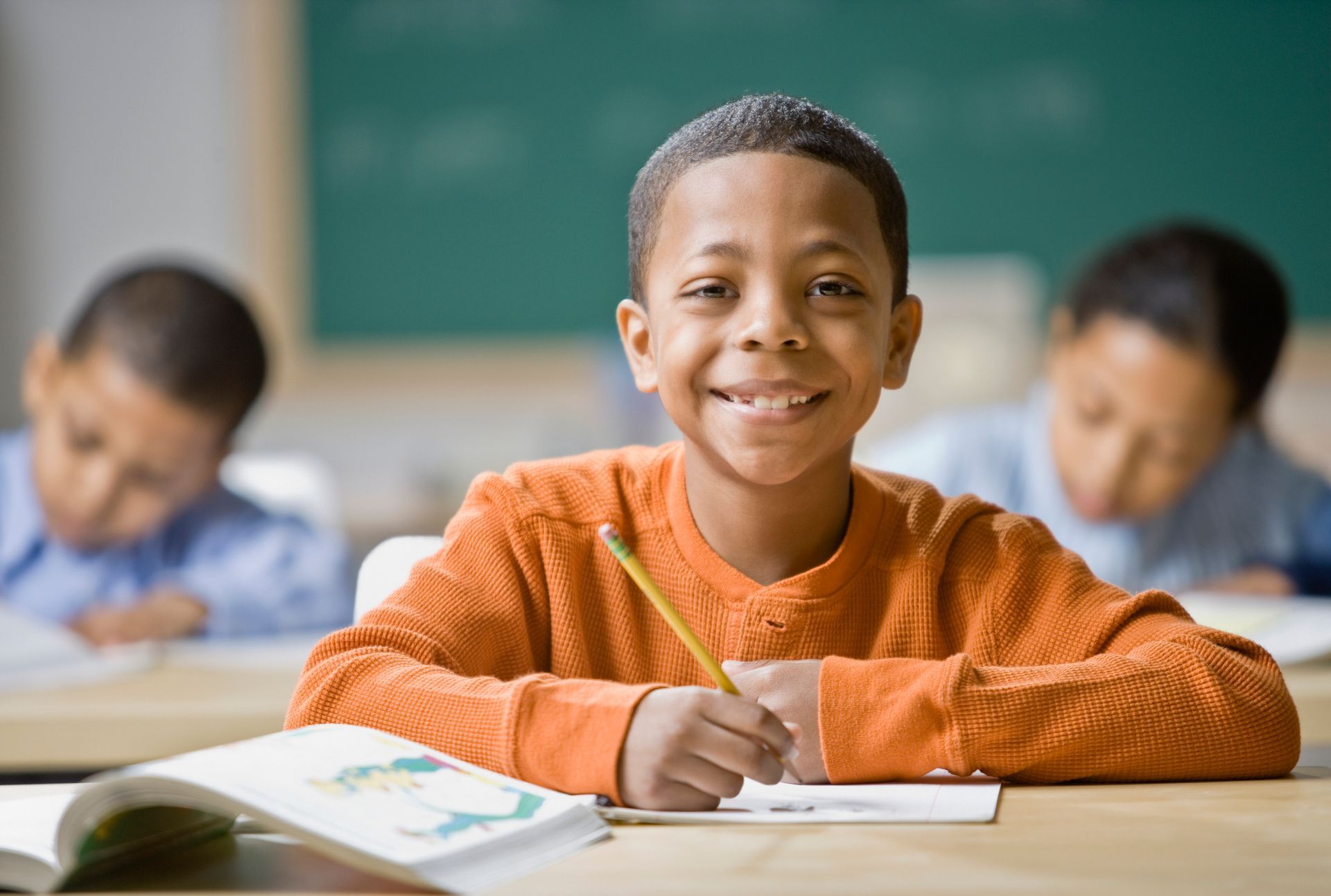 A young boy is smiling while sitting at a desk in a classroom.