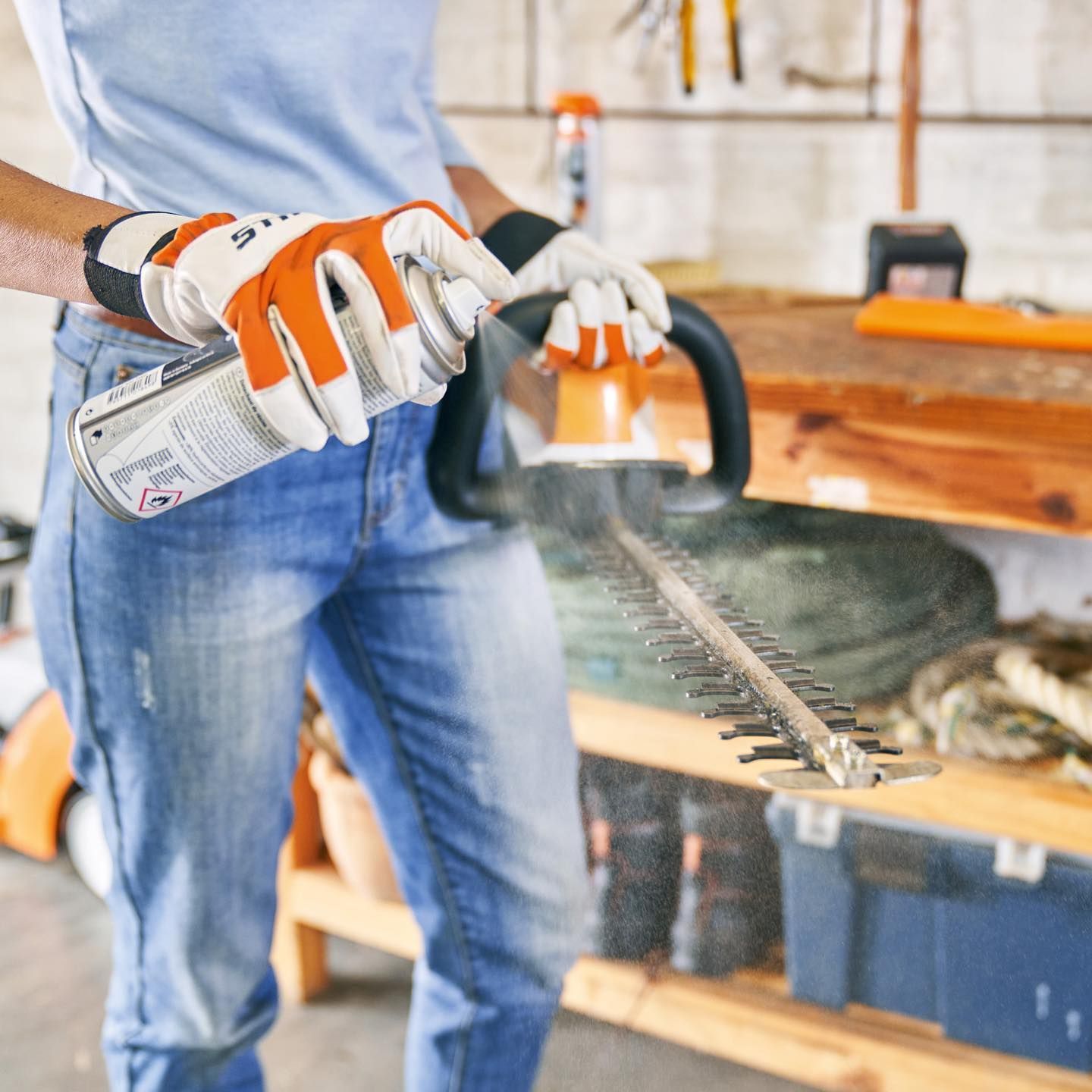 Person Sprays Lubricant On Hedge Trimmer Blade In A Garage, Wearing Gloves And Jeans — Stihl Shop Atherton In Atherton, QLD