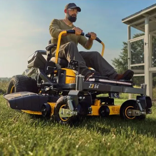 Man riding a yellow zero-turn lawn mower through tall grass beside a house— Stihl Shop Atherton In Atherton, QLD