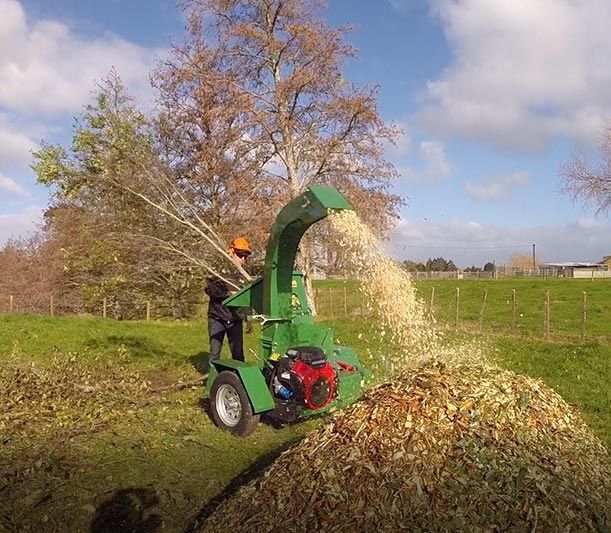 A green wood chipper blows shredded branches into a pile in a grassy field.— Stihl Shop Atherton In Atherton, QLD