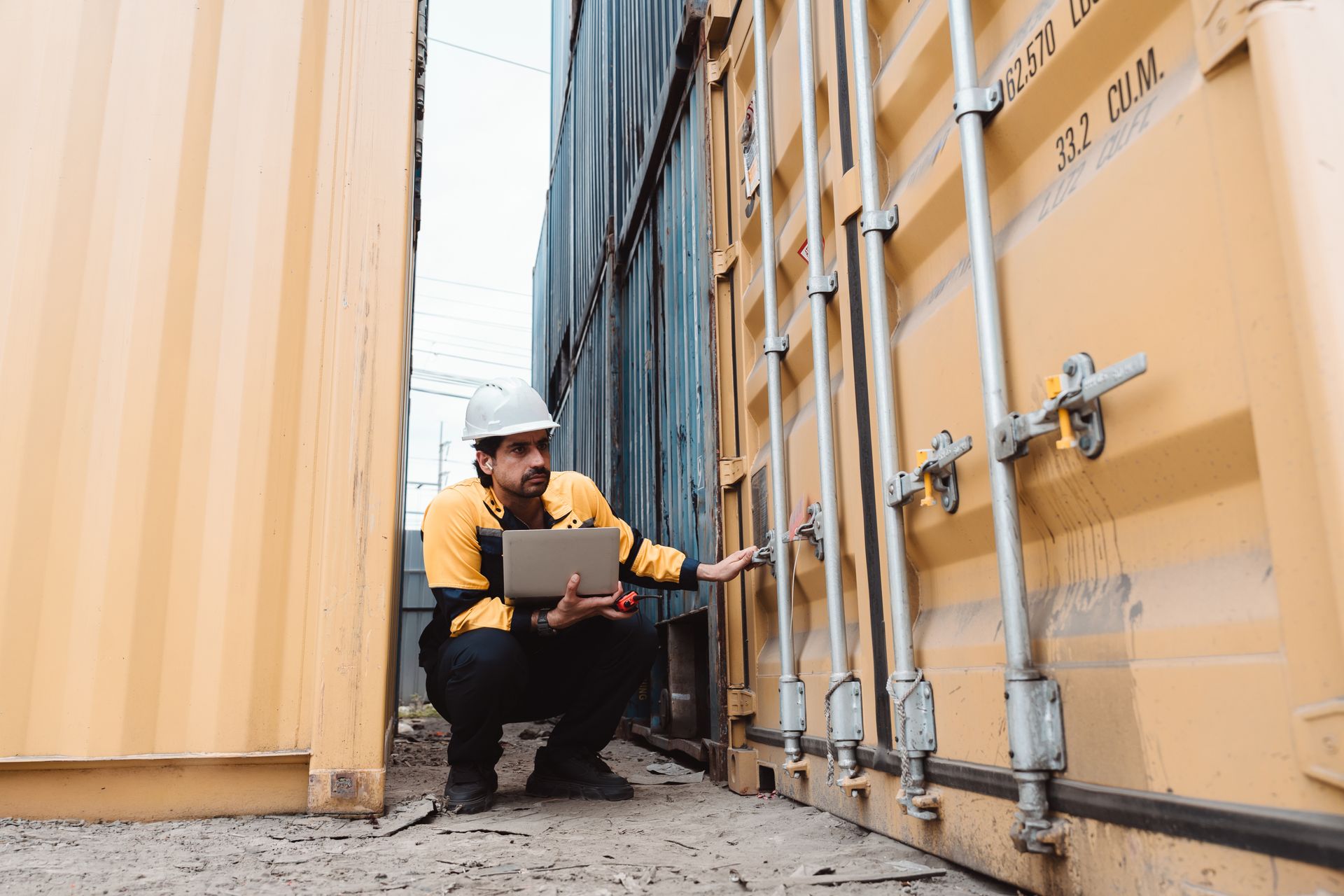 Industrial door supplier technician inspecting shipping container doors with laptop on job site.