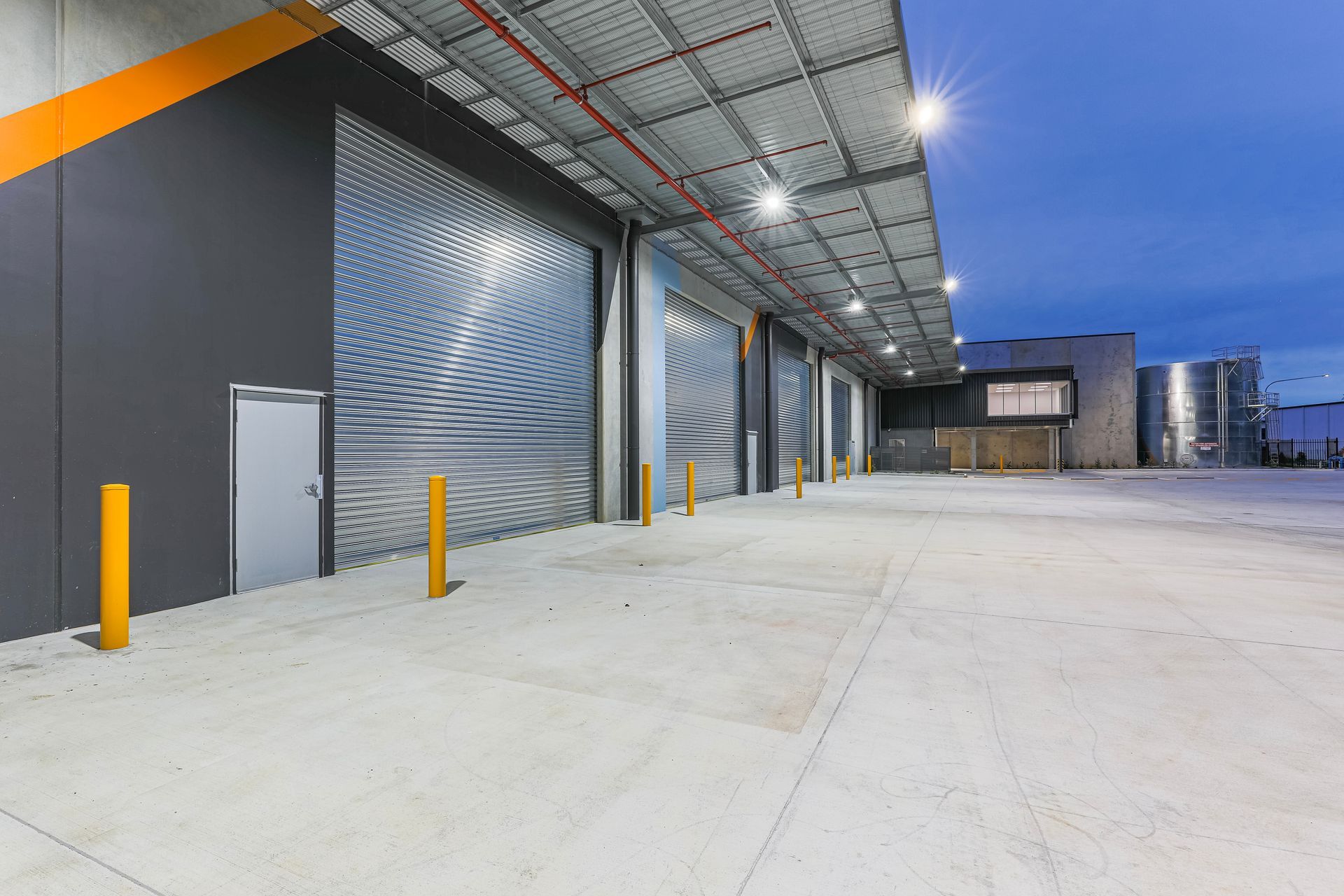 An industrial warehouse exterior at dusk, featuring multiple large gray metal roll-up doors and a concrete loading area.