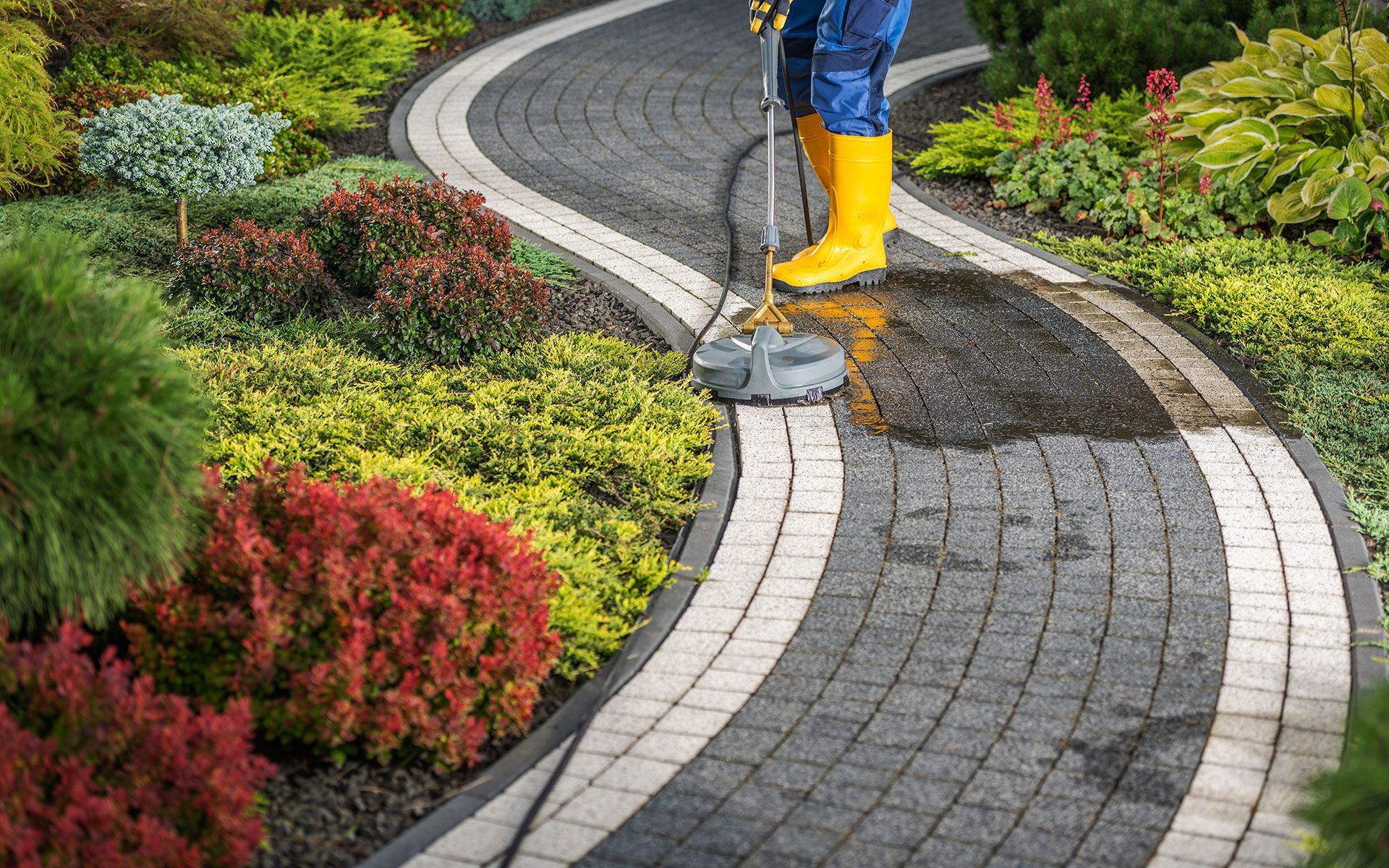 A man in yellow boots is cleaning a brick walkway in a garden.