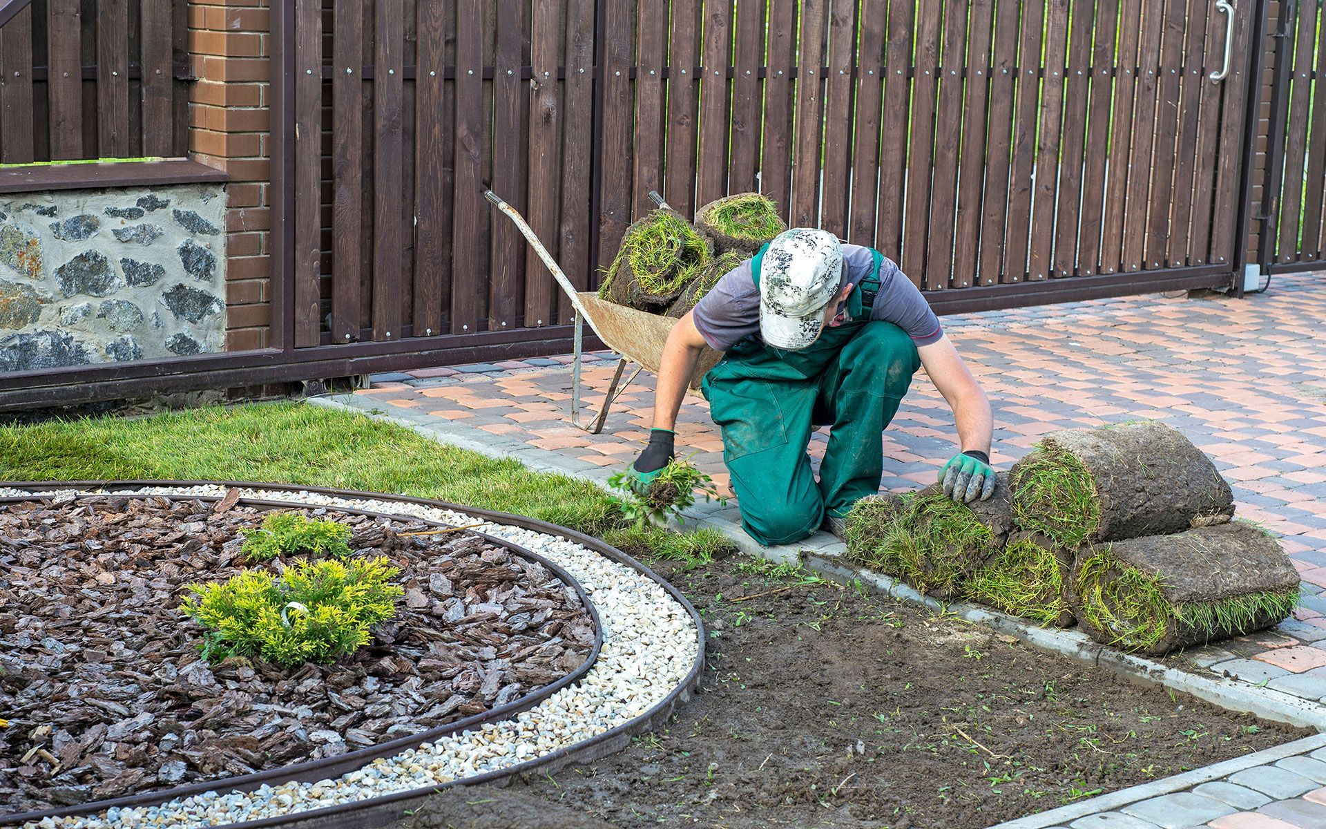 A man is kneeling down in a garden with a wheelbarrow.