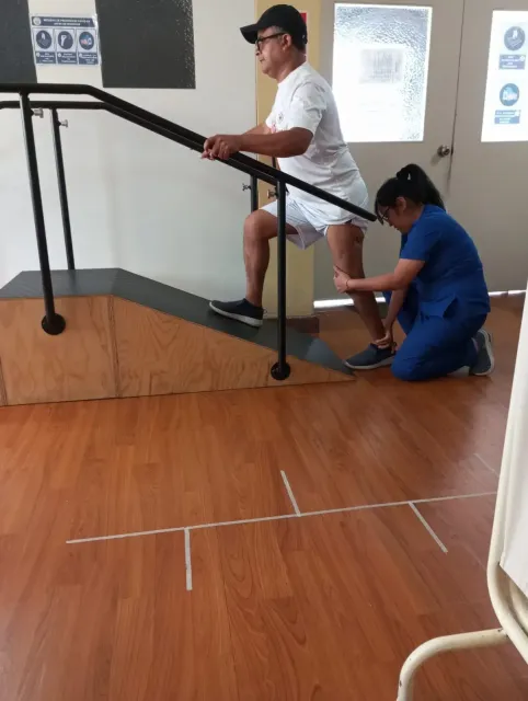 Un hombre con camisa blanca usa una rampa mientras otra persona estabiliza su andador en una habitación con suelo de madera
