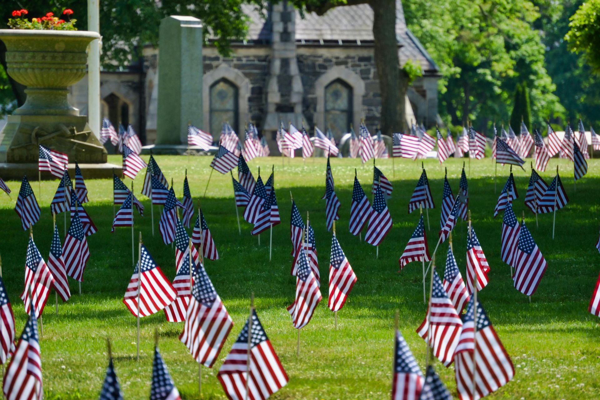 Rows of small American flags on a grassy cemetery lawn with gravestones in the background.
