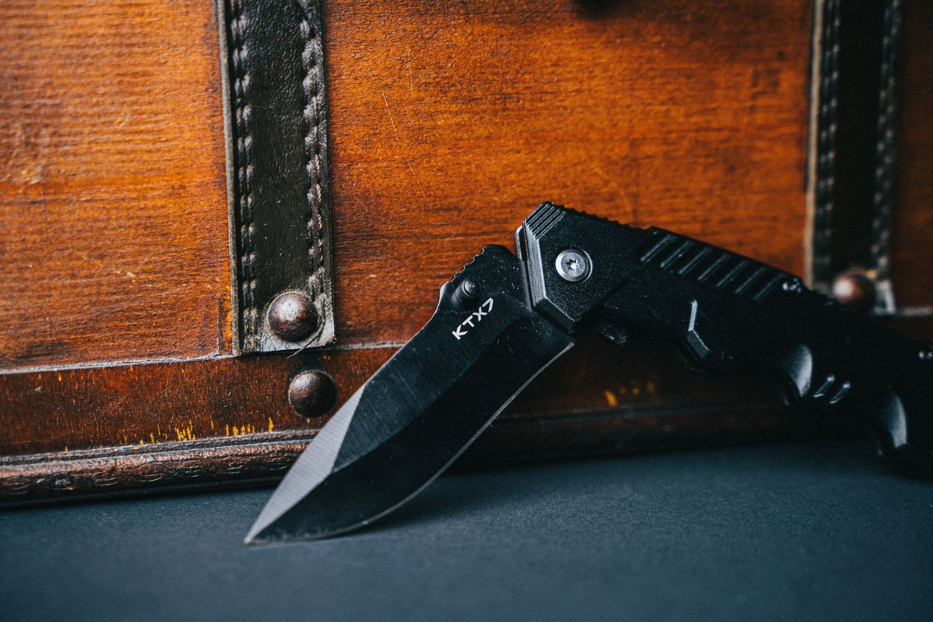 Black folding knife leaning against a brown leather suitcase on a dark surface